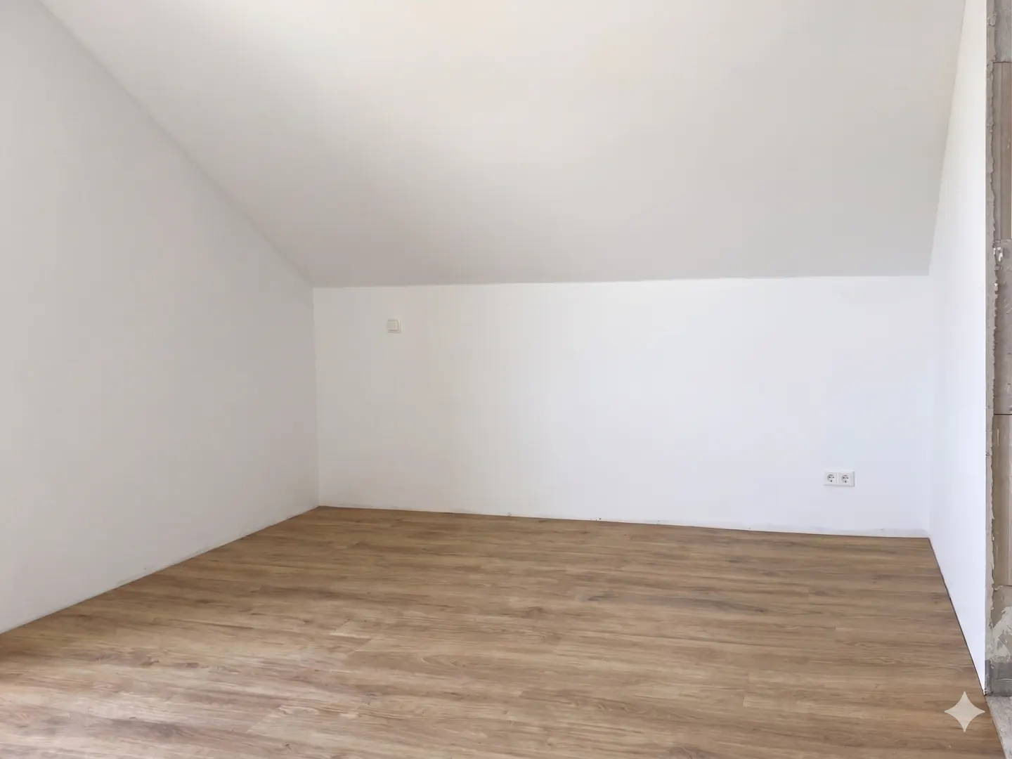 Empty room with white walls, sloped ceiling, and light brown wood flooring. Electrical outlets are visible on the right wall.