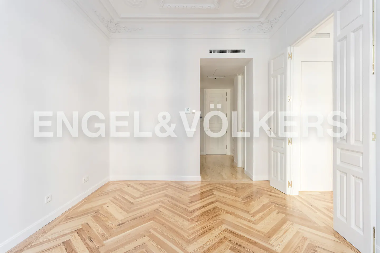 Bright, empty room with white walls, herringbone wood floor, and open white double doors leading to a hallway. Ceiling has decorative molding.