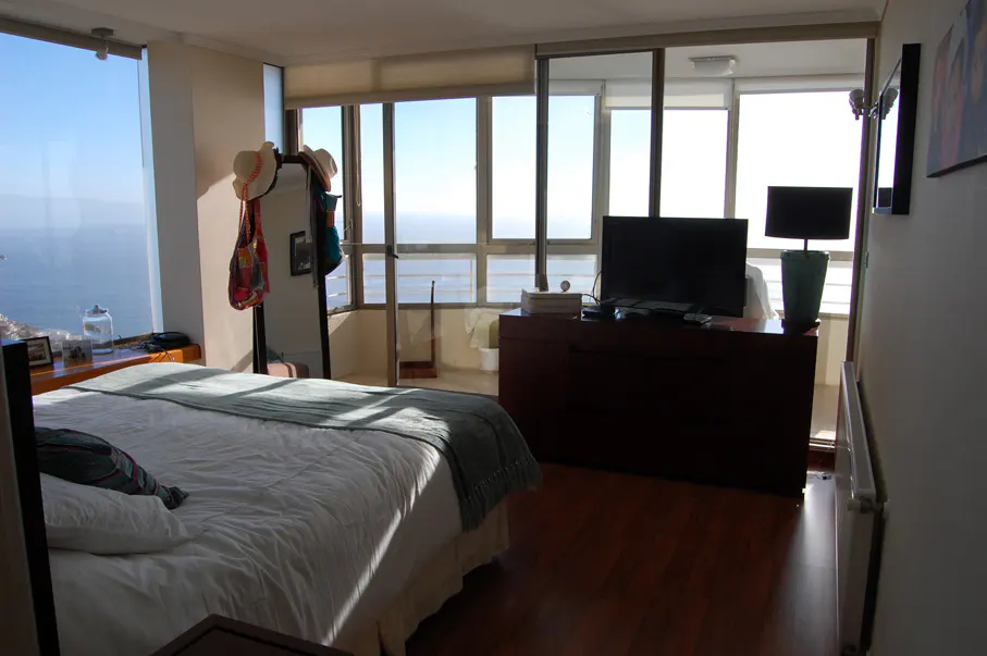 Bright bedroom with ocean view. White bed, dark wood dresser with TV, and floor-to-ceiling windows. Hats hang on a rack.