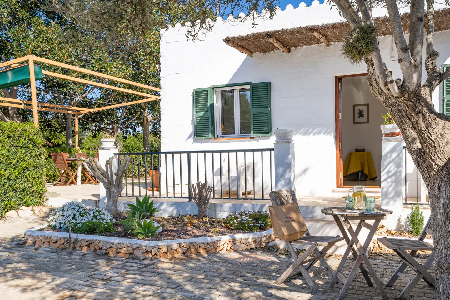 Exterior view of a white house with green shutters, a table with drinks, and a garden with flowers.