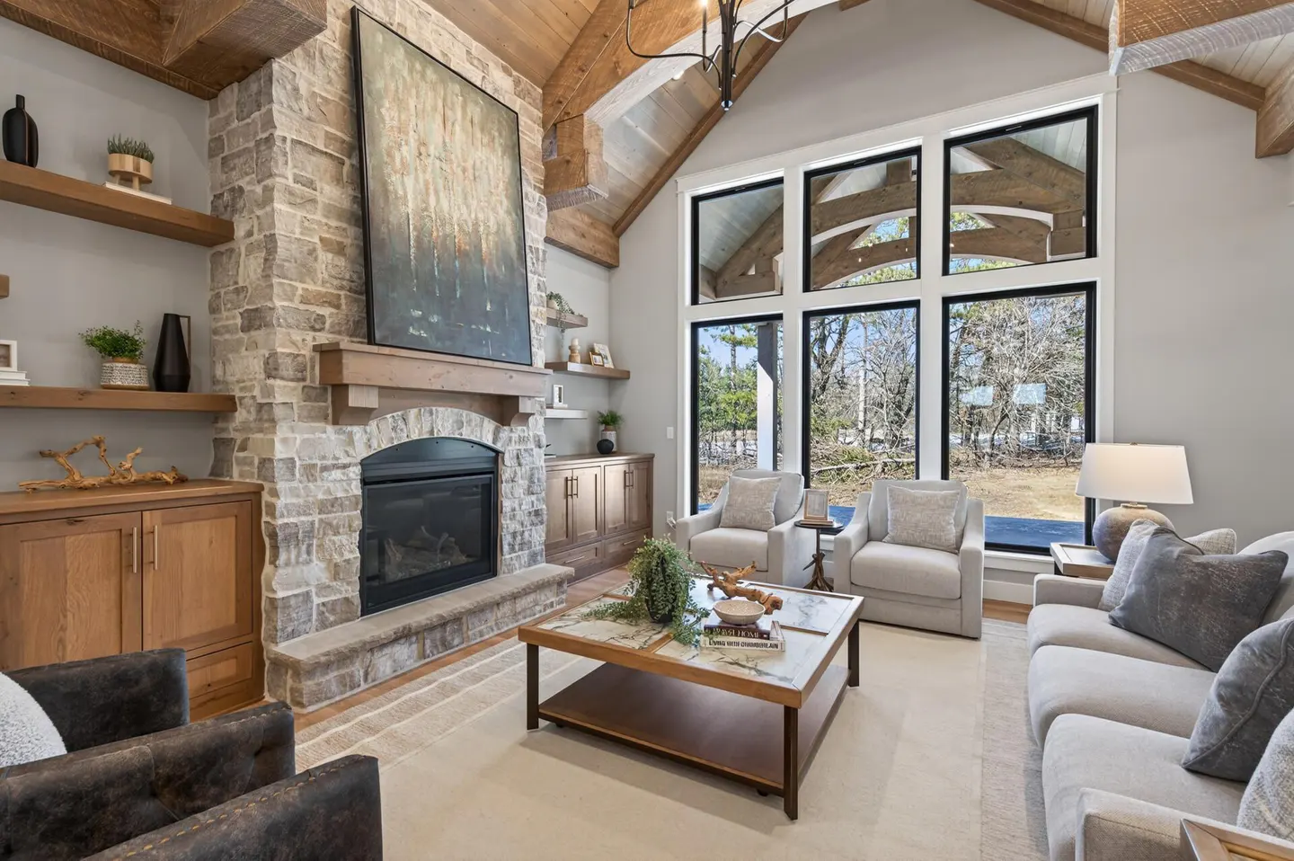 Living room with stone fireplace, wood beams, and large windows. Neutral colors, modern decor, and a view of trees outside.