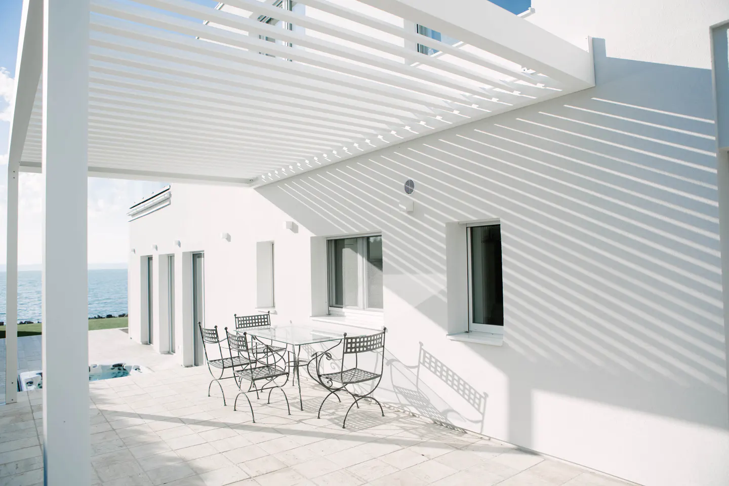 White modern house with a patio, table and chairs. The patio has a pergola, casting shadows on the wall. Ocean view in the background.