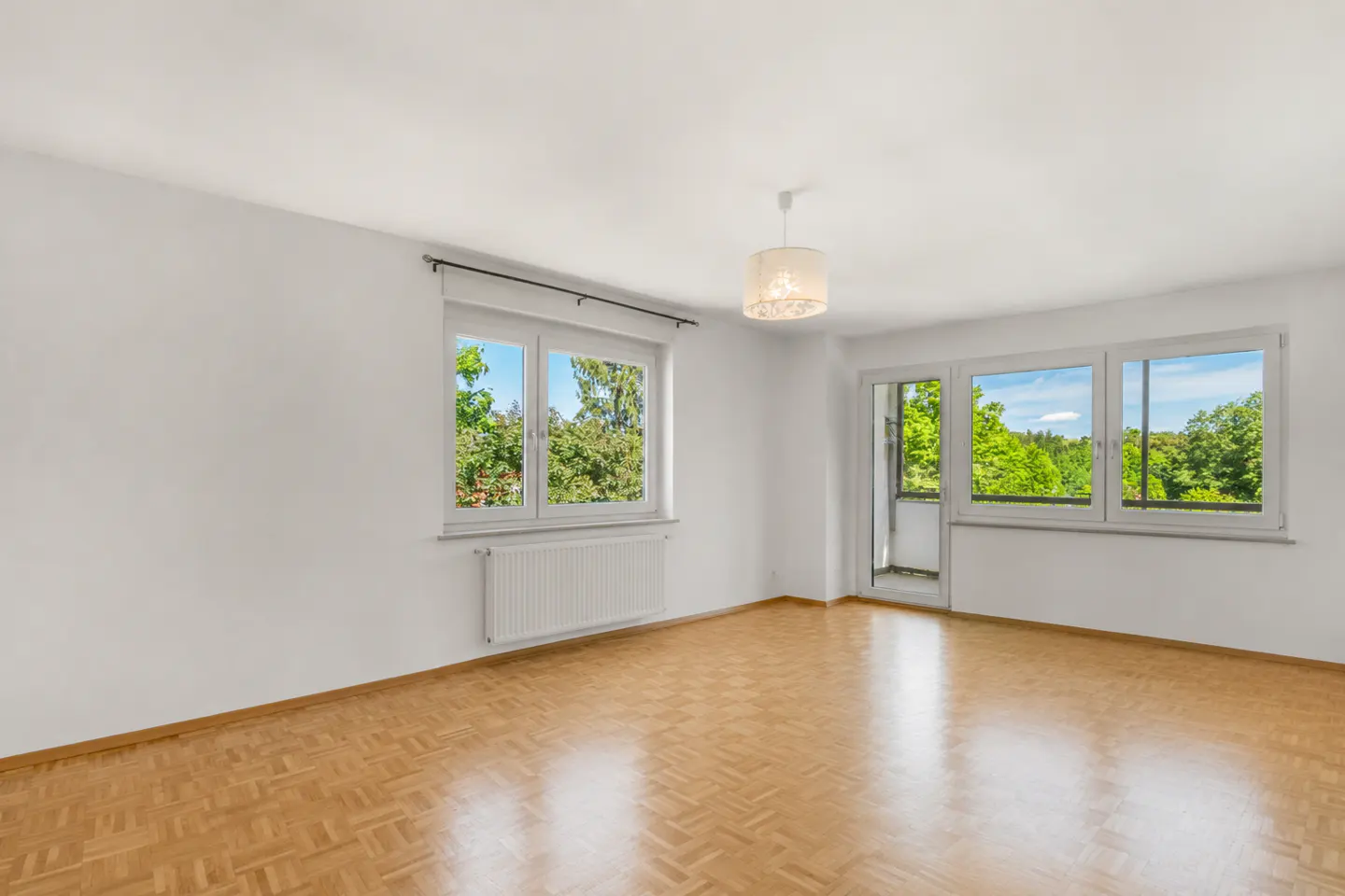 Bright, empty room with parquet floors, white walls, and two windows showing green trees. A white radiator is under one window. A light fixture hangs from the ceiling.
