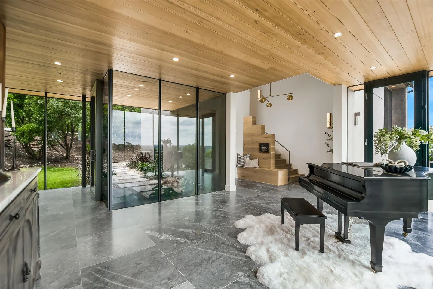 A modern home interior with a black grand piano, wooden stairs, and floor-to-ceiling windows overlooking a green landscape.