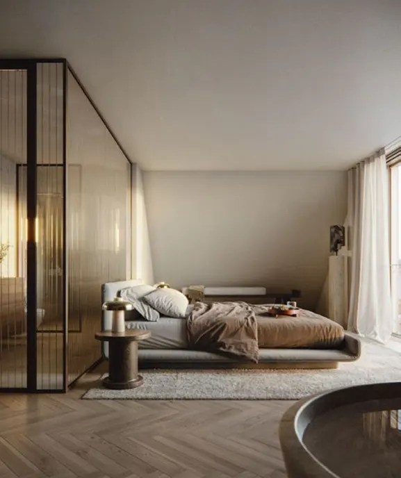A modern bedroom with a low platform bed, brown duvet, and a round side table with a lamp. A glass-enclosed bathroom is visible.