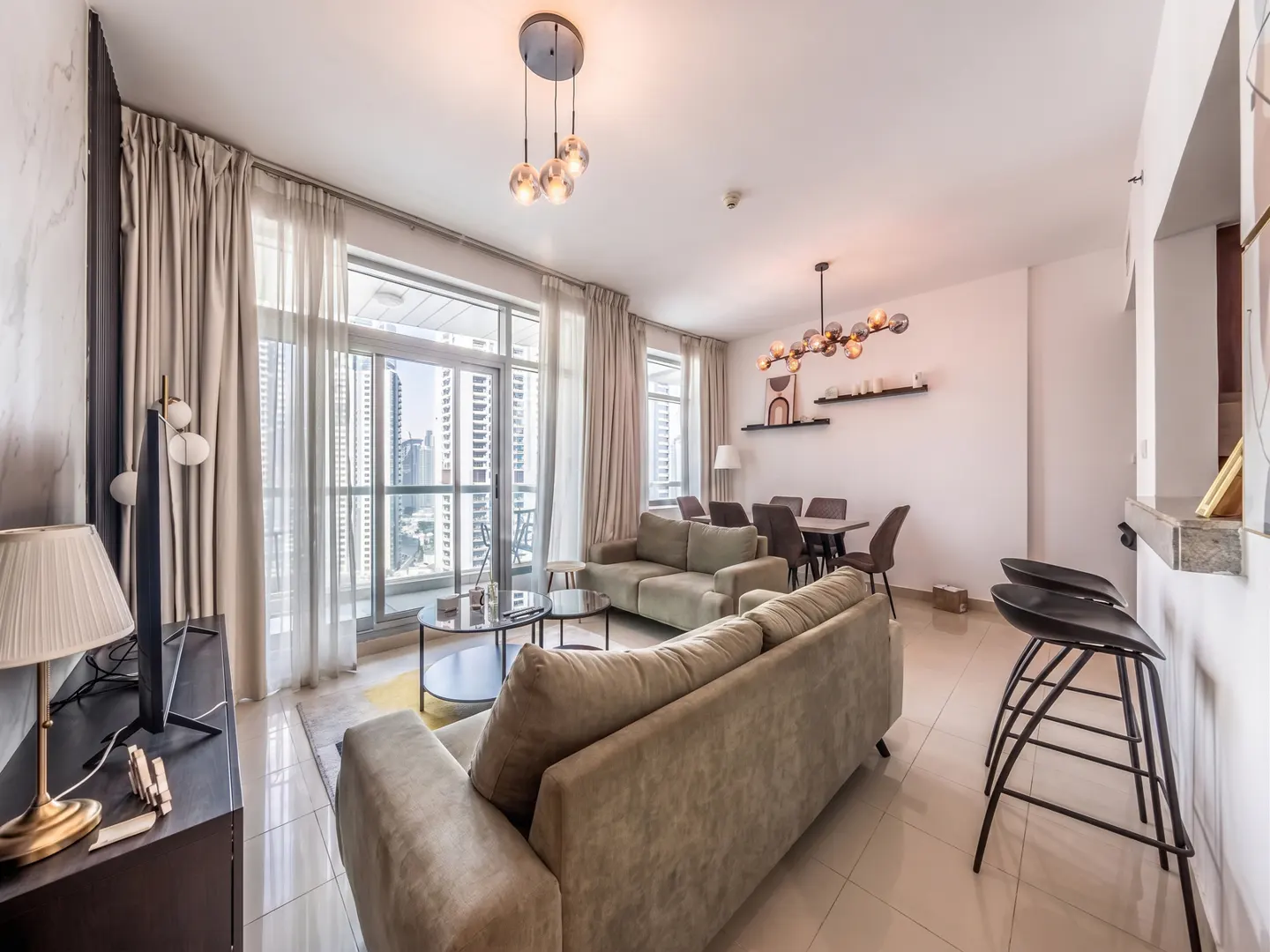 A bright, modern living room with beige sofas, a dining table, and a city view through large windows.