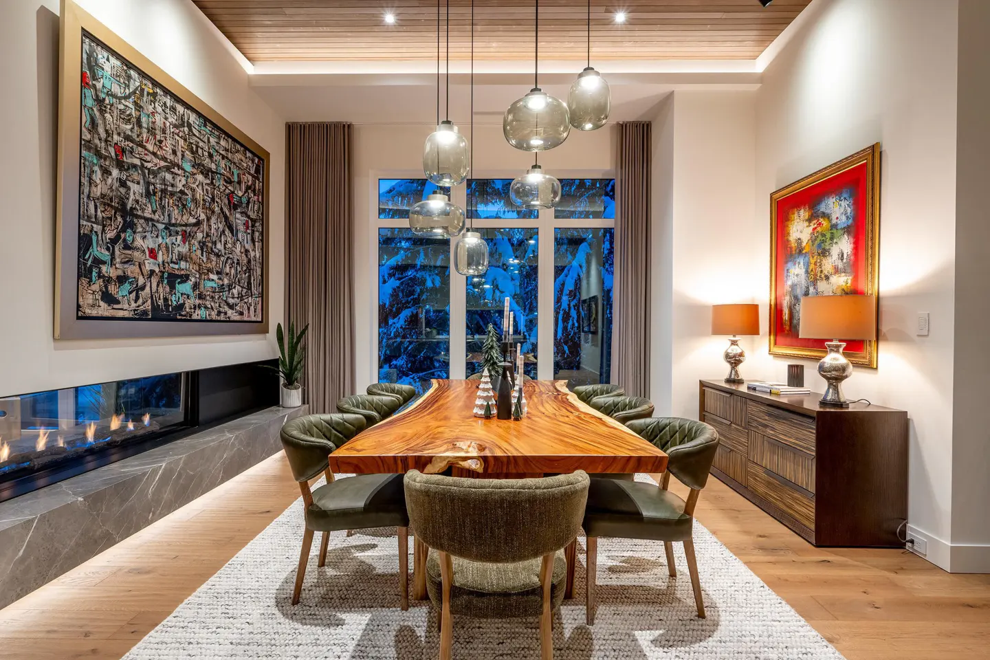 A dining room with a live-edge wood table, green chairs, and pendant lights. A fireplace and abstract art add to the room's ambiance.