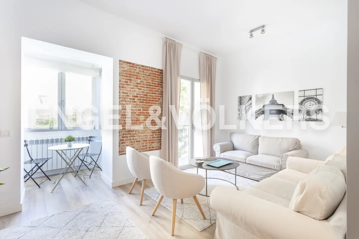 Bright living room with a brick accent wall, white sofas, two armchairs, a coffee table, and a small balcony dining area.