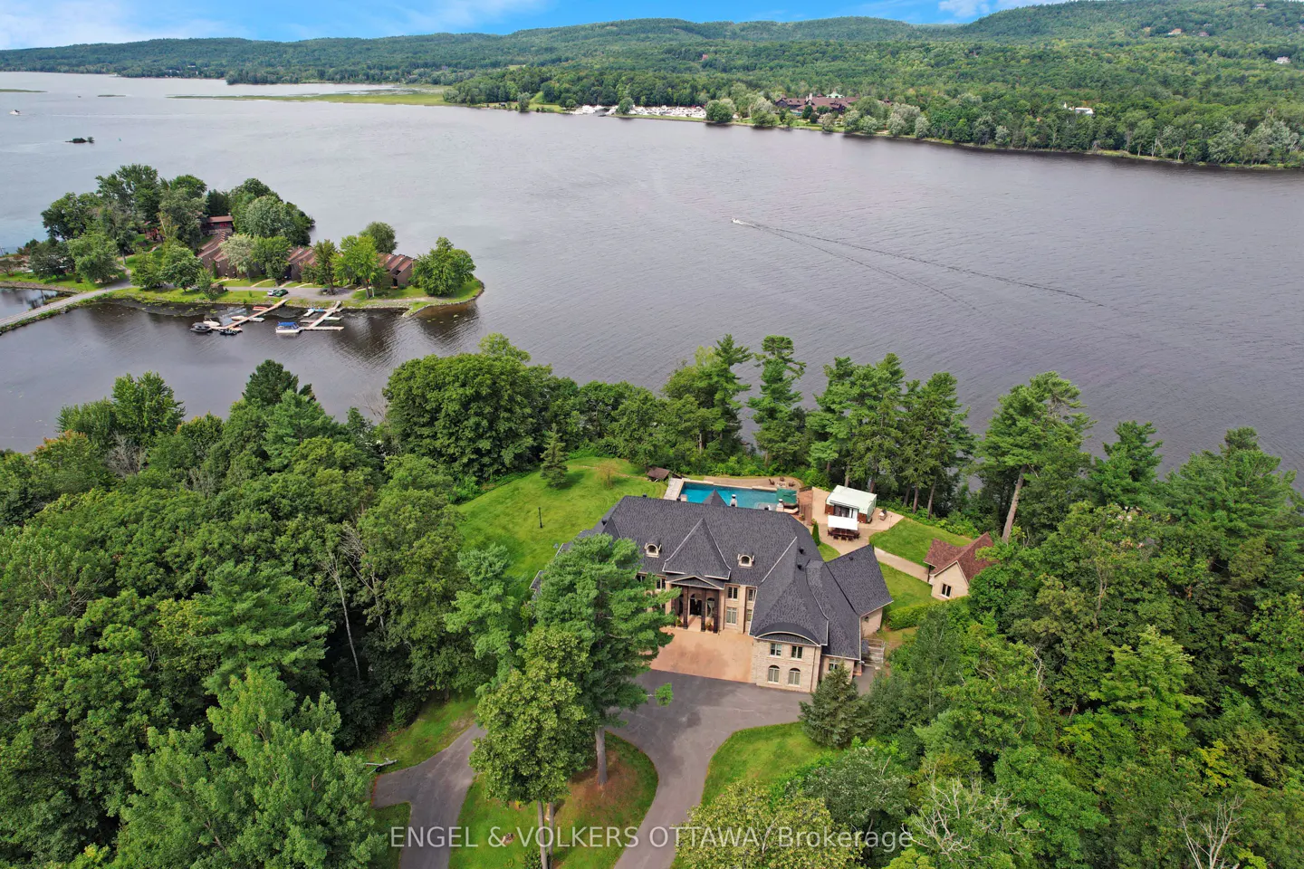 Aerial view of a large tan house with a black roof, pool, and lake in the background.
