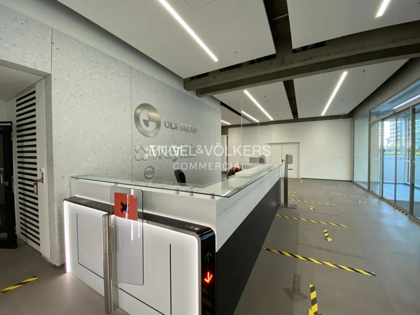 Modern office lobby with a white reception desk, glass partition, and Engel & Völkers logo on a textured wall. Black and yellow floor markings.