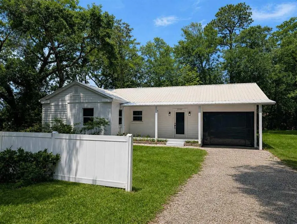 A white, one-story house with a black garage door and gravel driveway. A white fence is in the front yard.