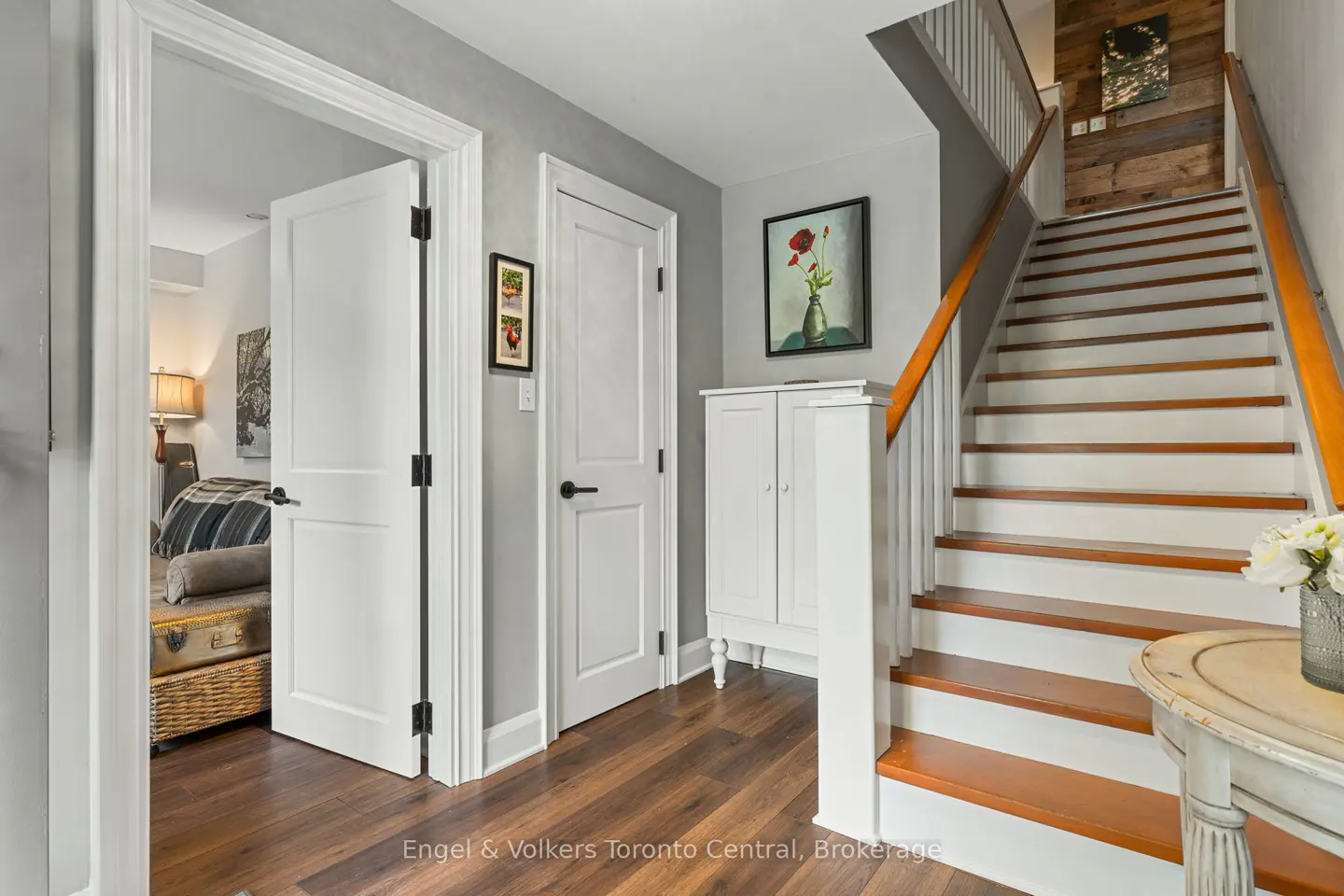 A home's foyer with hardwood floors, white doors, and a staircase with wood treads and white risers. A white cabinet and a table with flowers are also visible.