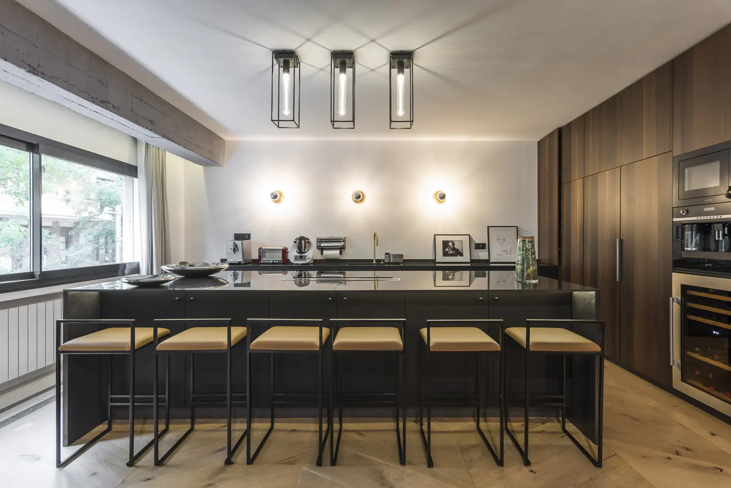 Modern kitchen with black island, tan stools, and wood floors. Stainless steel appliances line the back wall. Three pendant lights hang above.