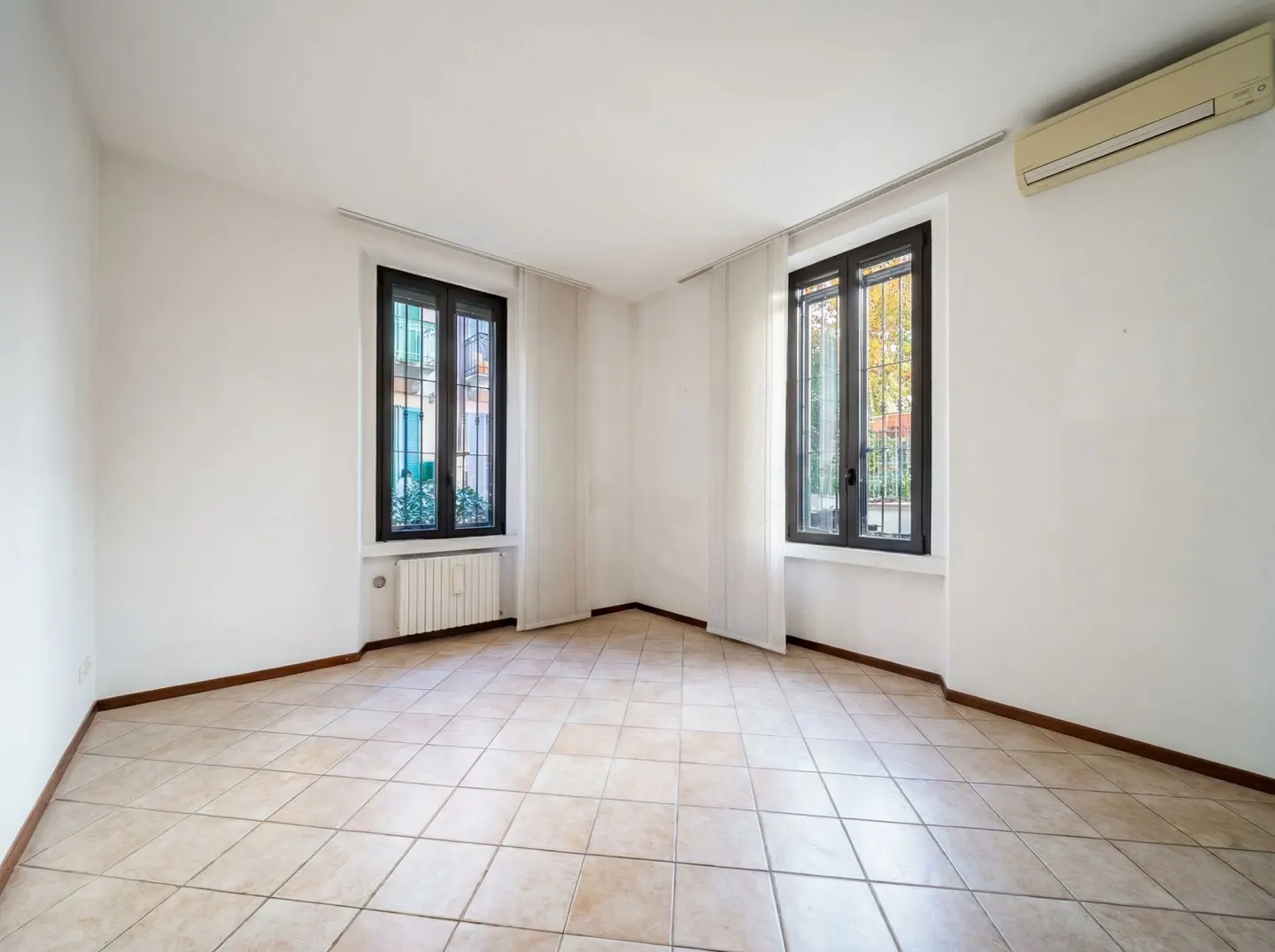 Empty room with white walls, beige tile floor, and two black-framed windows with white curtains. An air conditioner is mounted high on the wall.