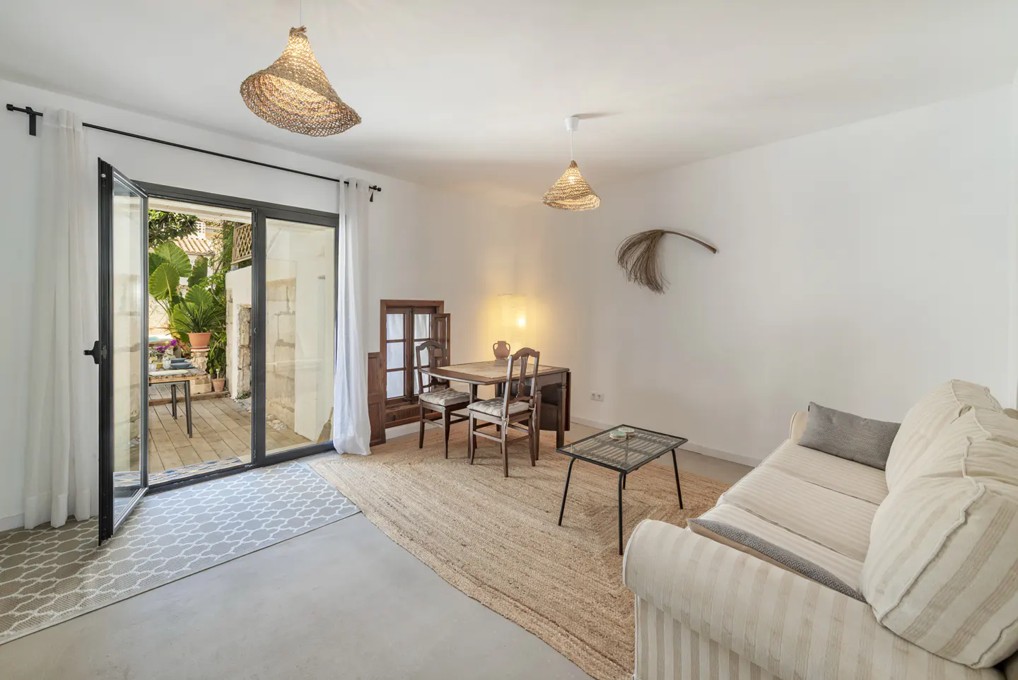 Bright living room with a sofa, table, and open patio doors leading to a garden. Woven lamps hang from the ceiling.