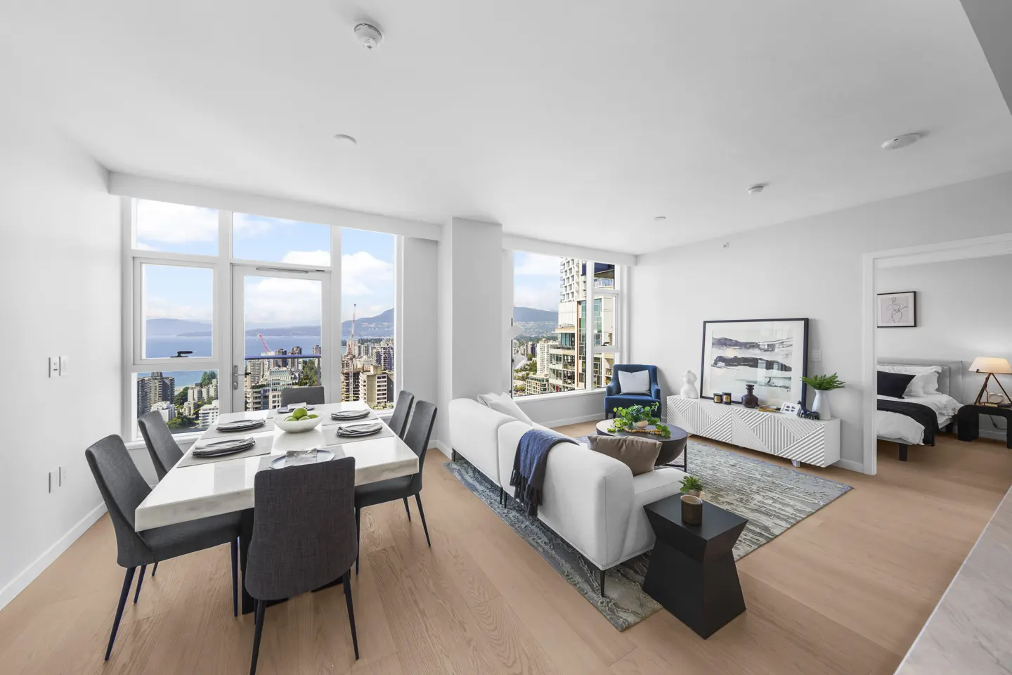 Bright, modern condo interior with dining table, white sofa, and city view. Bedroom visible through doorway.