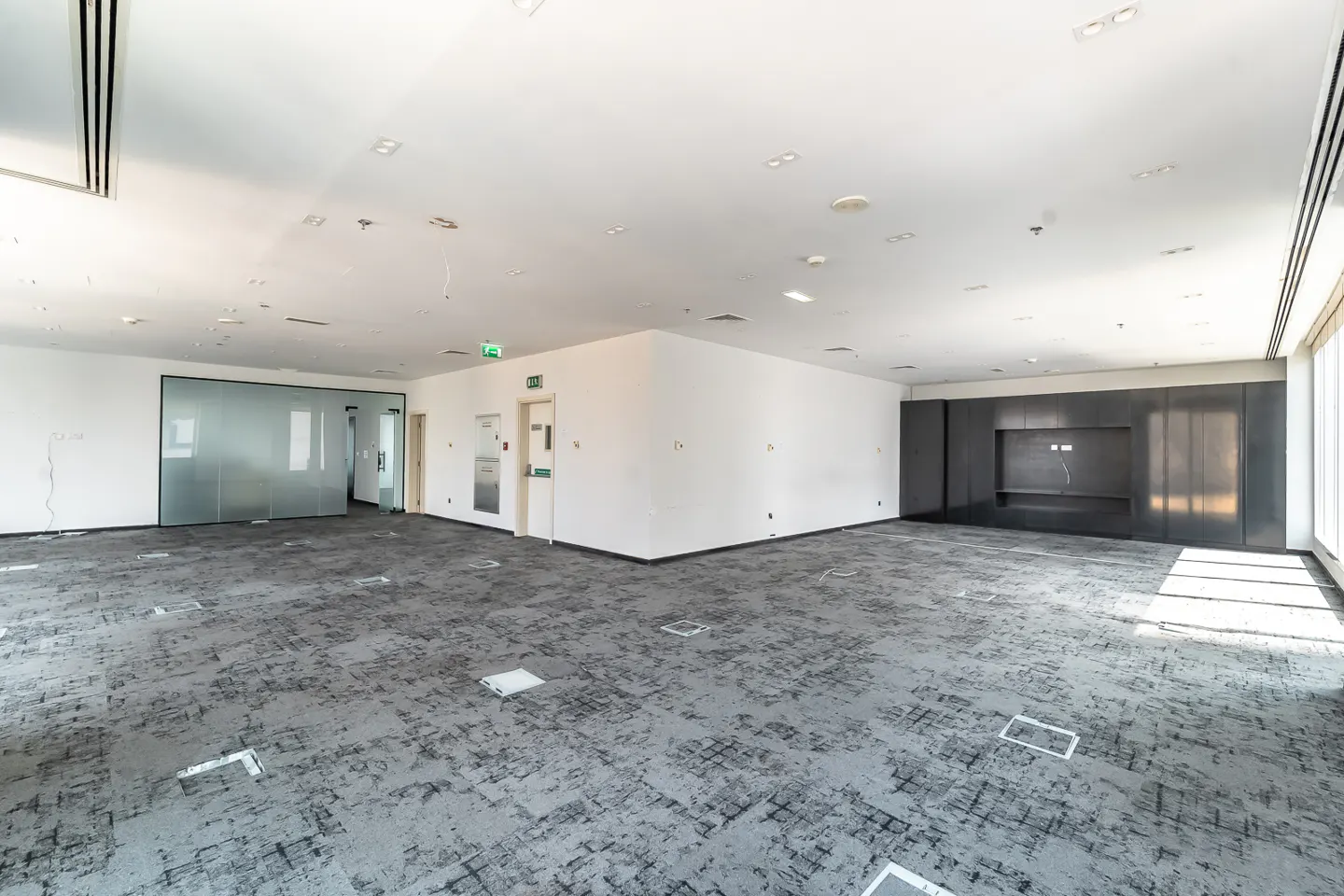 Bright, empty office space with gray patterned carpet, white walls, and a dark accent wall. Windows provide natural light.