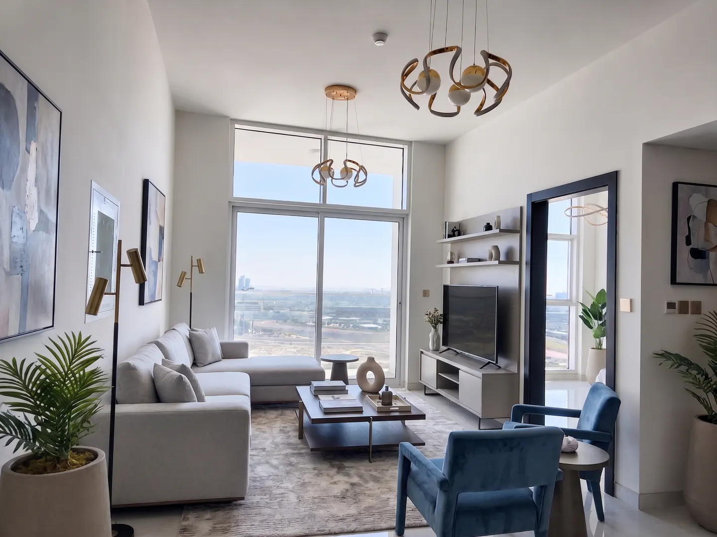 Bright living room with white walls, a gray sectional sofa, blue armchairs, and a large window with a city view. Modern light fixtures hang from the ceiling.