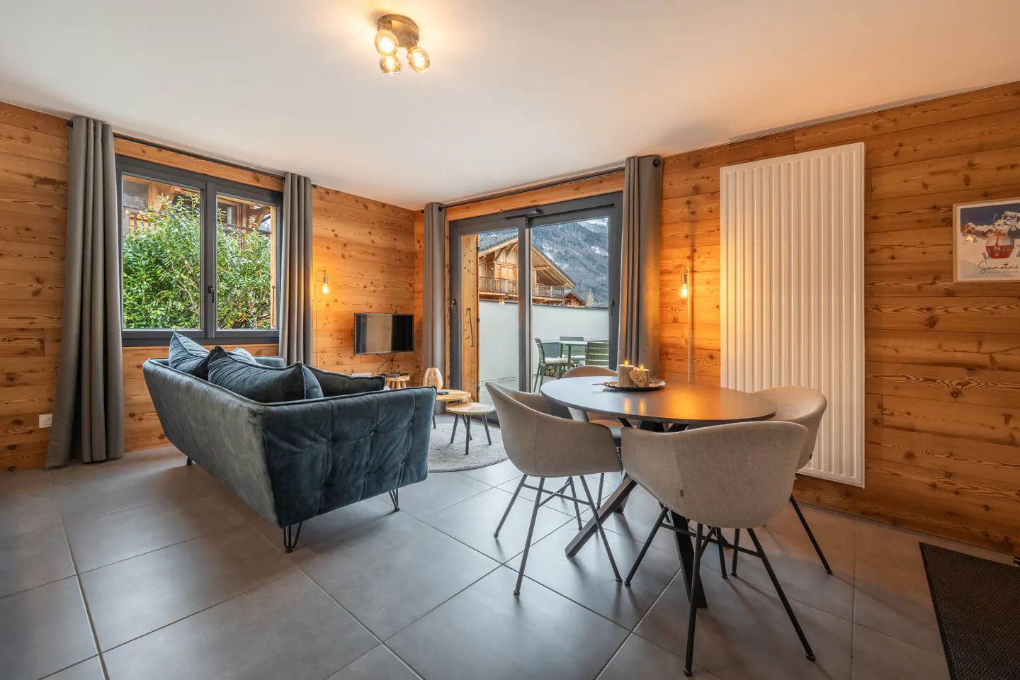 Living room with wood paneled walls, gray tile floor, blue sofa, and round dining table with four chairs. Balcony with mountain view.