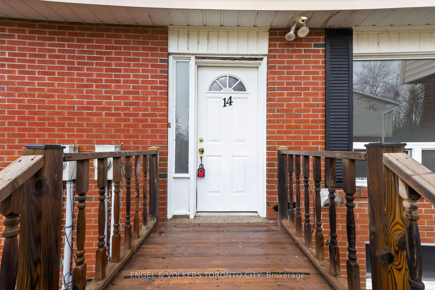 Exterior view of a red brick house with a white door numbered 14, accessed by a wooden ramp with railings.