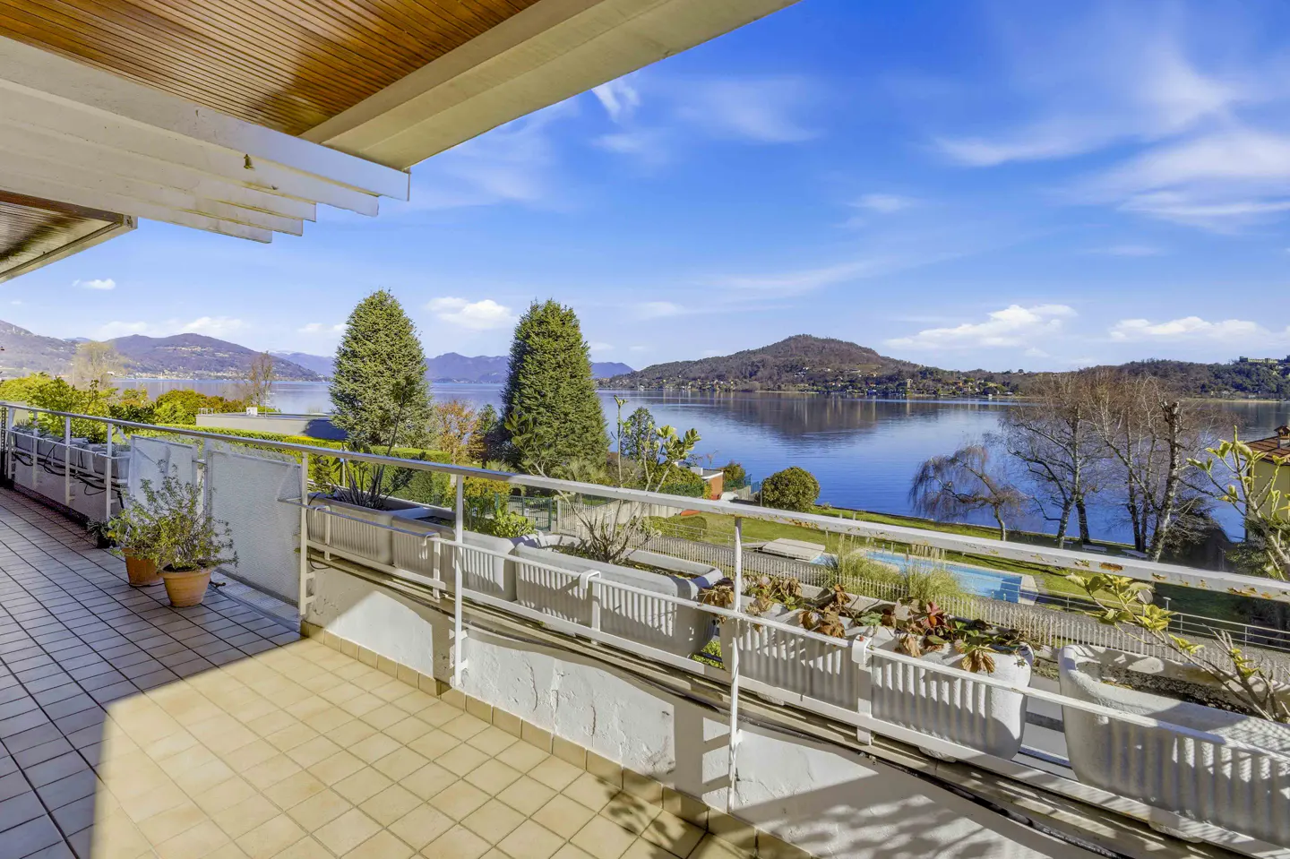 Balcony view of a blue lake with mountains in the background on a sunny day. Railing with plants and tiled floor.