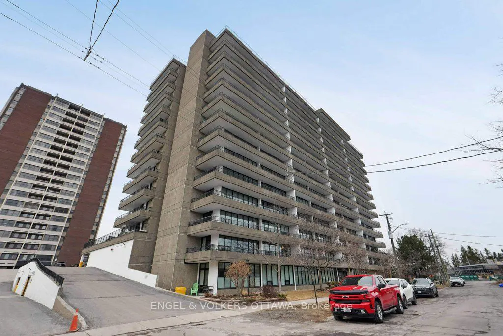 Exterior view of a tall, gray apartment building with balconies, cars parked on the street, and another building in the background.