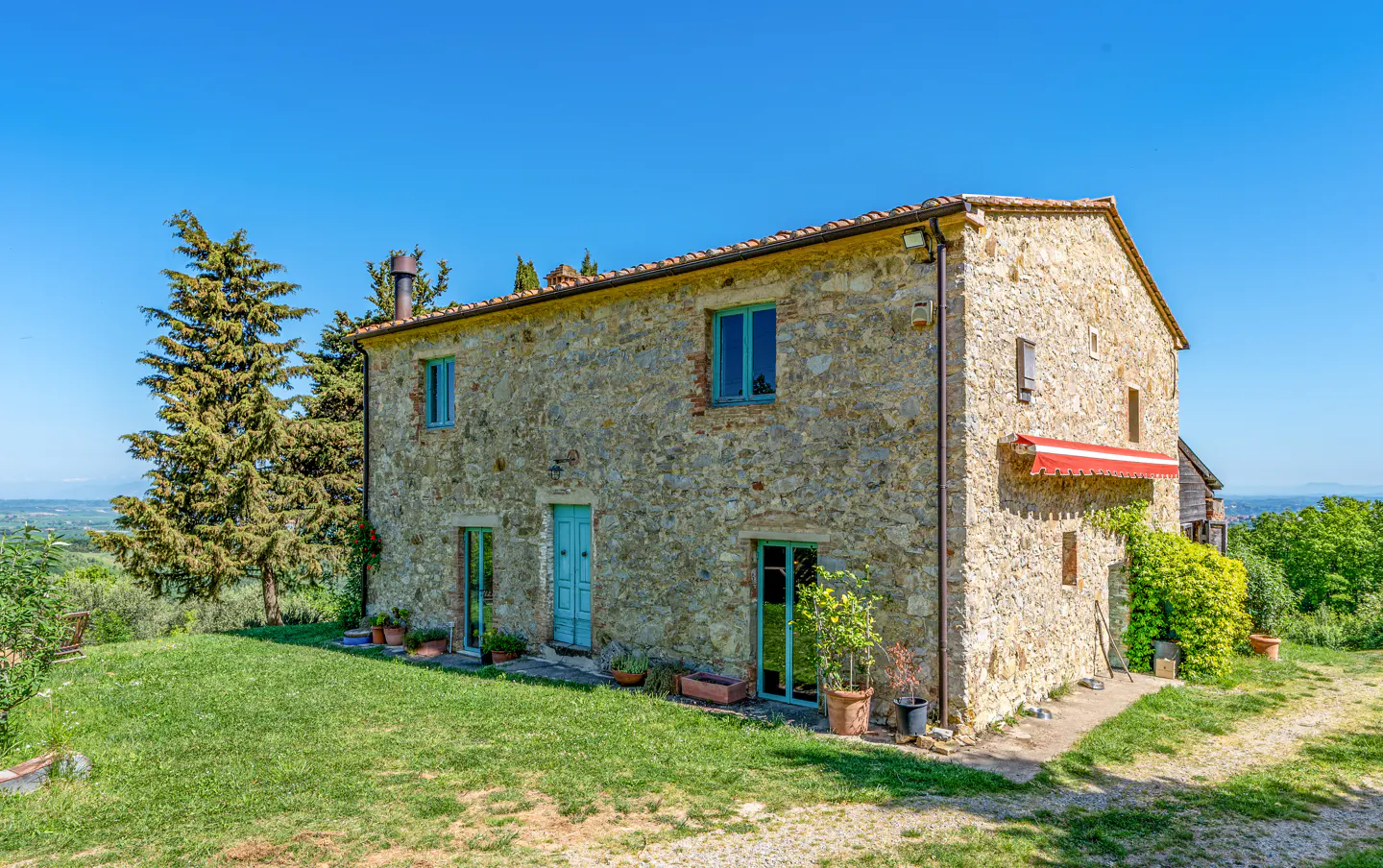 Exterior view of a two-story stone house with blue trim, a red awning, and a green lawn under a clear blue sky.