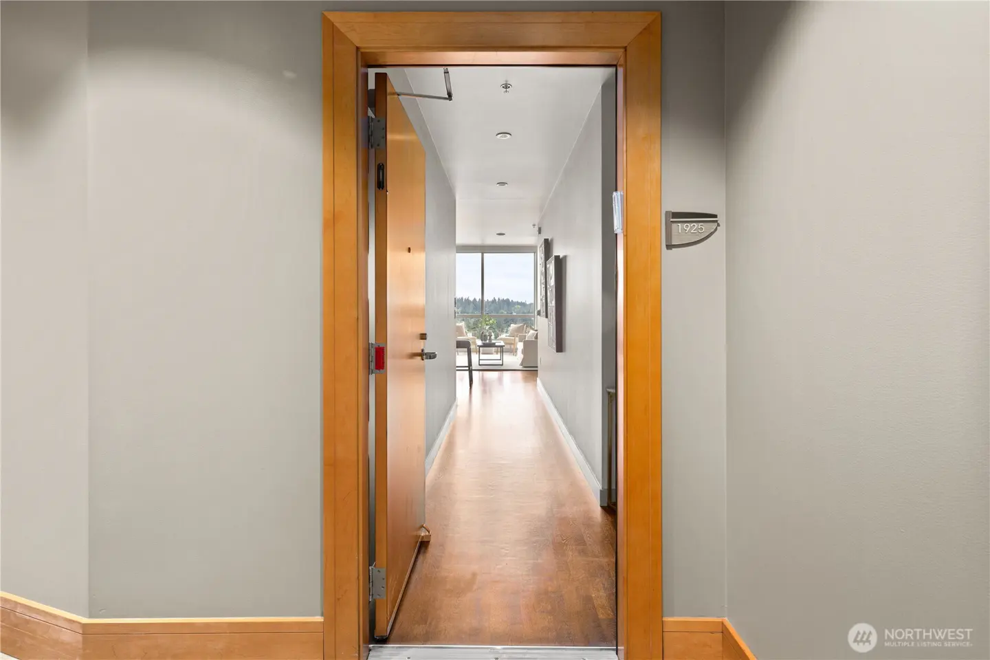 Open doorway to a condo hallway with wood floors, gray walls, and a view of a living room and city skyline.
