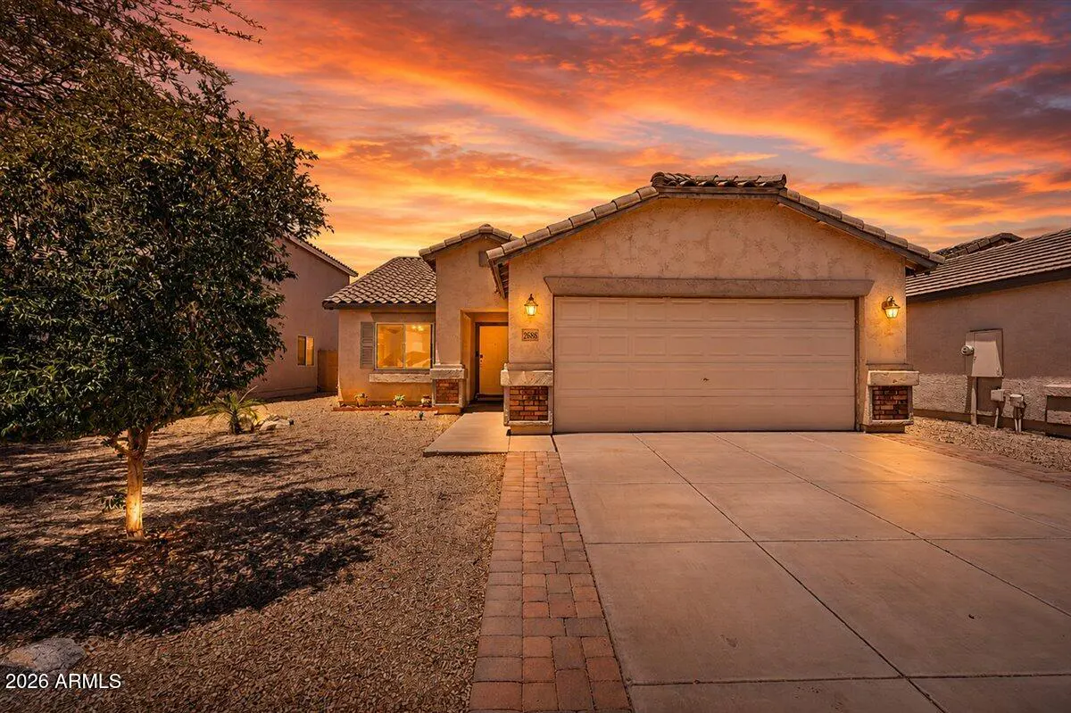 Single-story stucco home with a two-car garage and brick-trimmed driveway under a vibrant orange sunset sky.