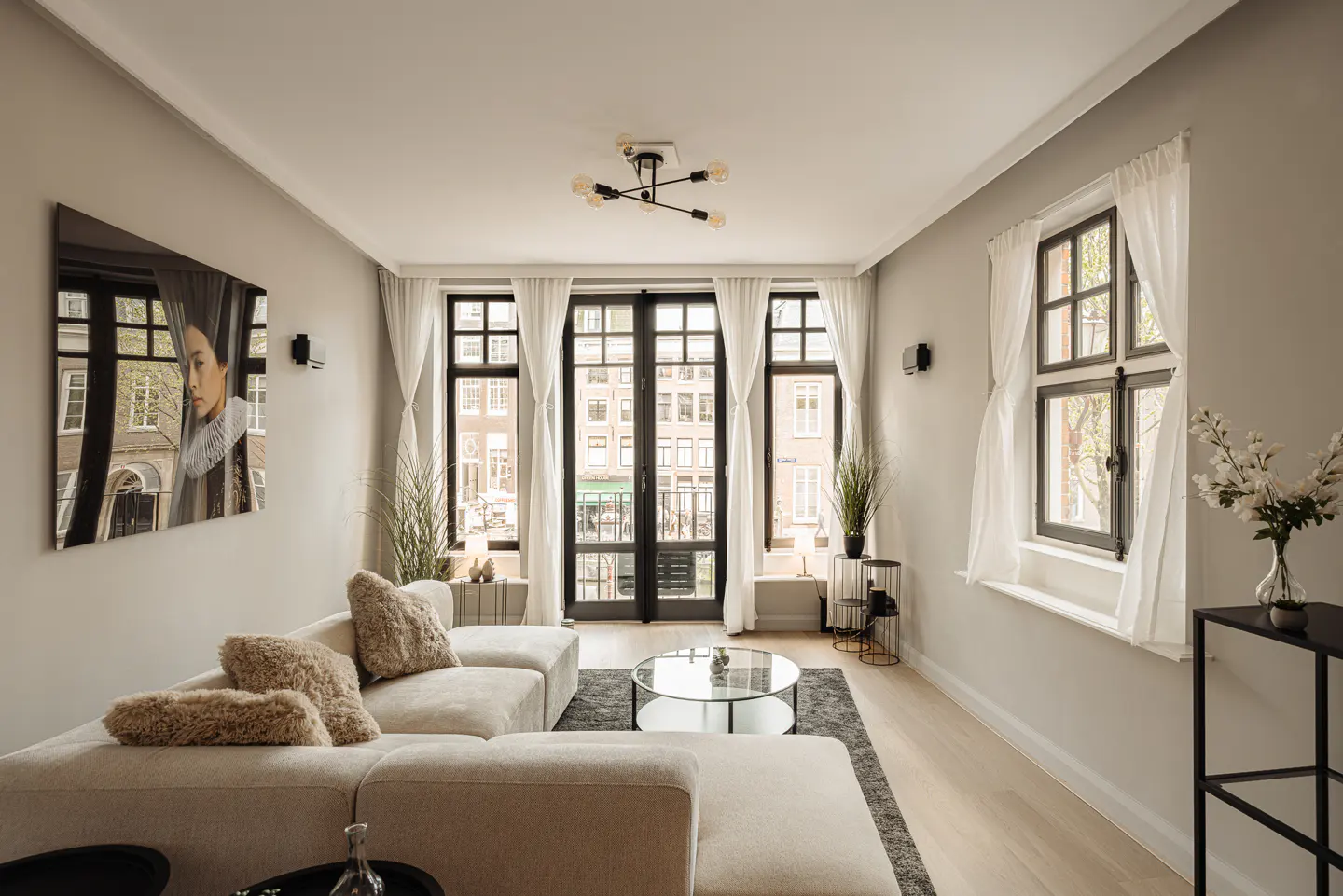 Bright living room with a cream sectional sofa, a glass coffee table, and black-framed windows with sheer white curtains. A large portrait hangs on the wall.