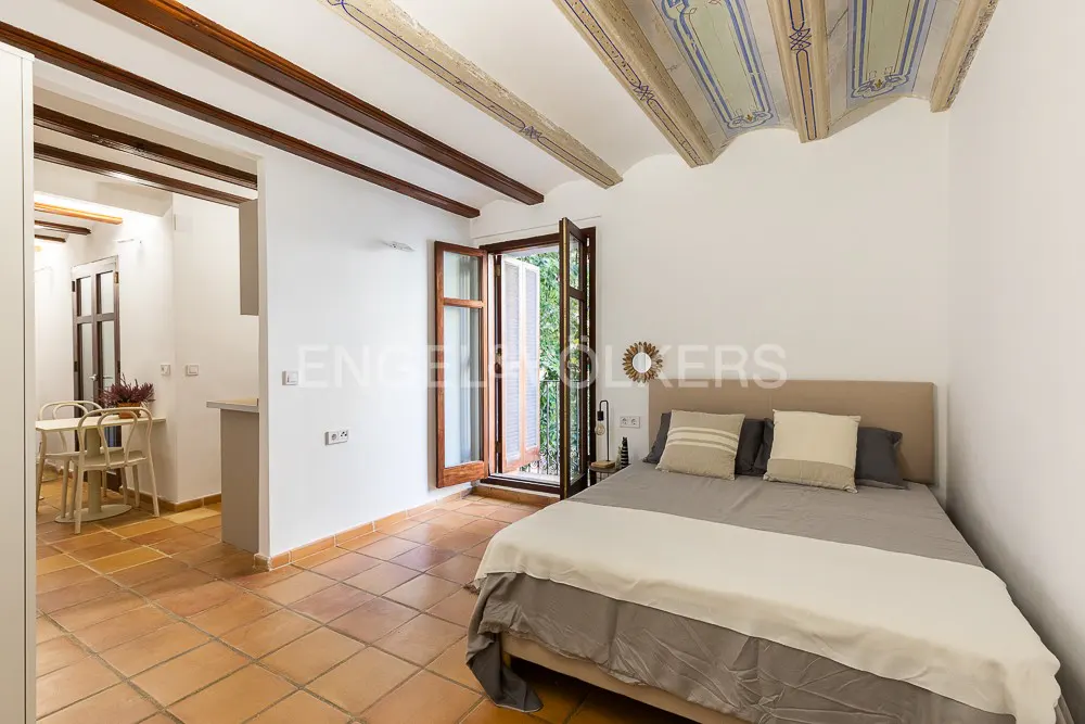 Bedroom with terracotta tile floor, white walls, wood beams, and bed with gray bedding. Open wood shutters lead to a balcony.
