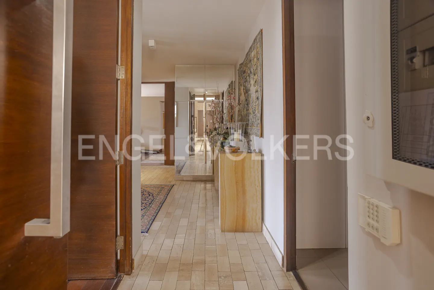A bright hallway with light-colored tile floors, a wooden console table, a wall tapestry, and a view into another room.