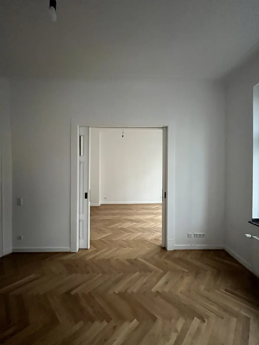 Empty room with white walls and light wood herringbone flooring, connected to an adjacent room by a wide doorway.