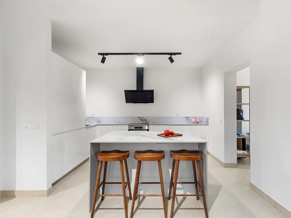 Bright, modern kitchen with white cabinets, marble countertops, and a central island with three wooden stools. Black range hood and track lighting above.