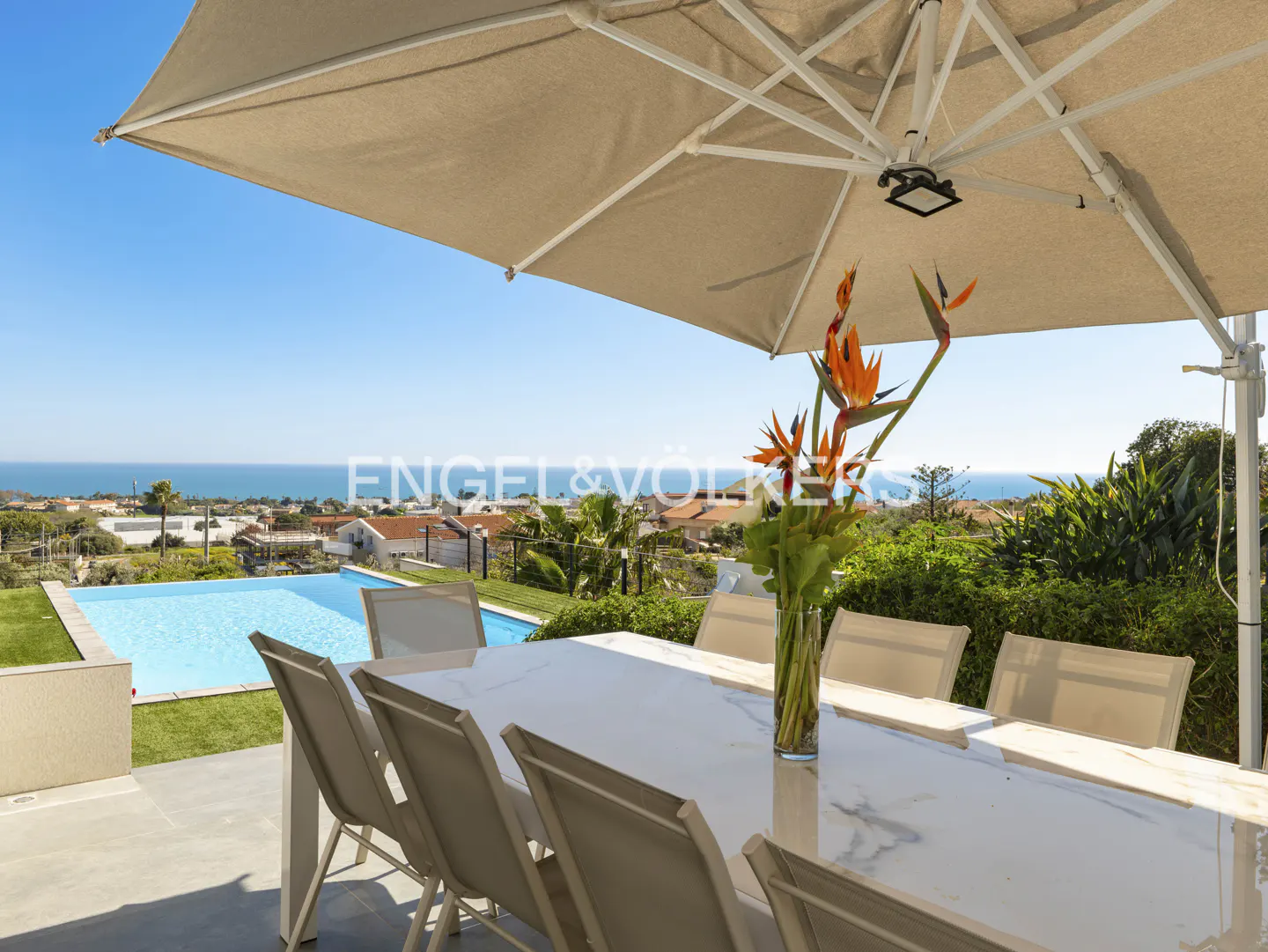 Outdoor dining area with a white marble table, chairs, and a vase of Bird of Paradise flowers, overlooking a pool, houses, and the ocean.