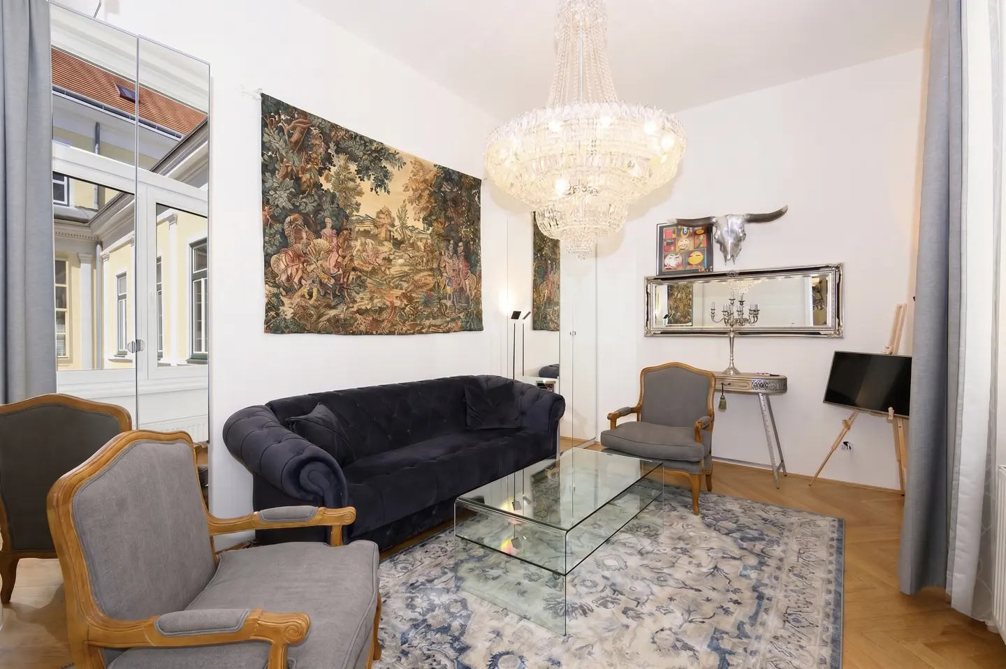 A living room with a blue sofa, two gray chairs, a glass table, and a large chandelier.