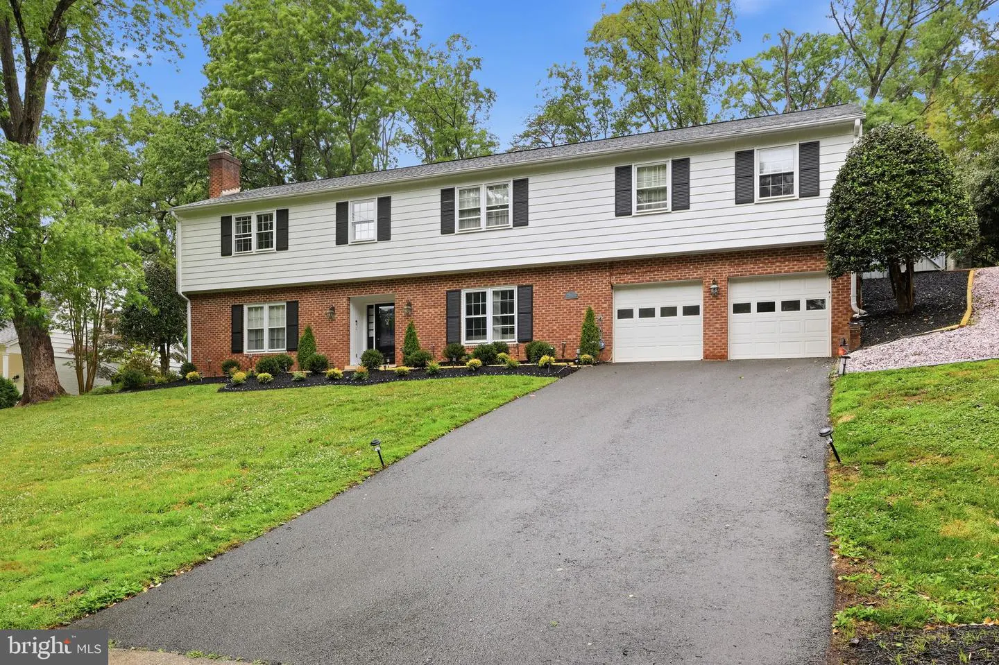 Two-story house with a brick and white facade, black shutters, and a two-car garage. A driveway leads up to the house.
