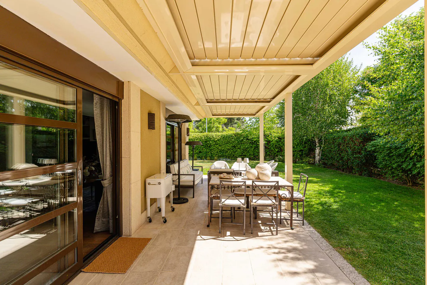 Covered patio with a dining table, chairs, and a white cooler. Green lawn and trees are visible in the background.