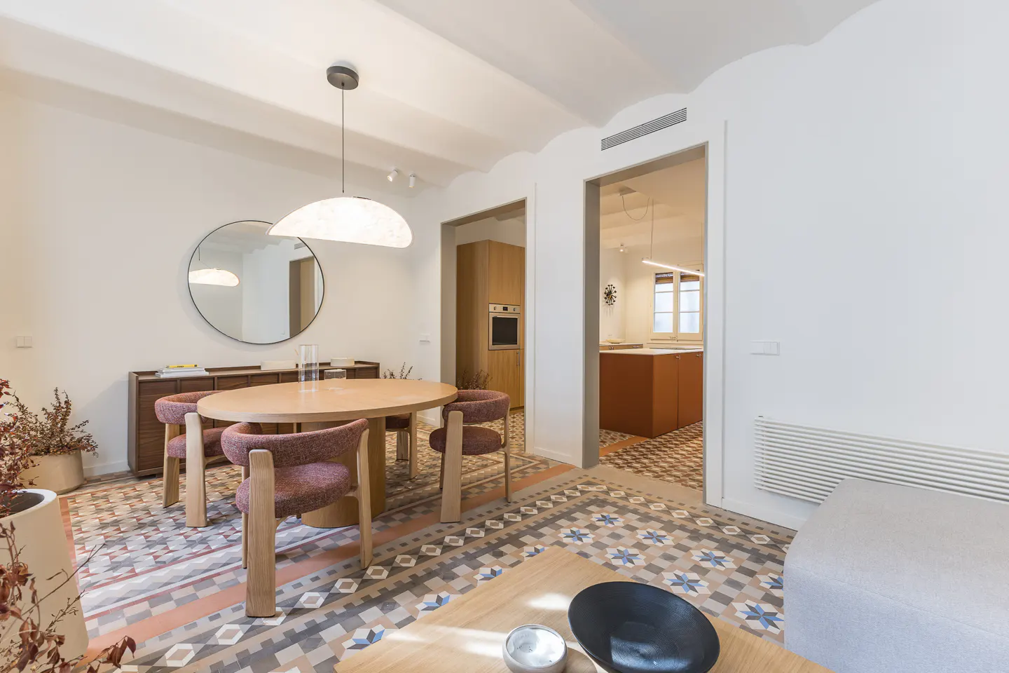 A bright, modern dining room with a round wooden table, four chairs, and patterned tile flooring. A kitchen is visible through a doorway.