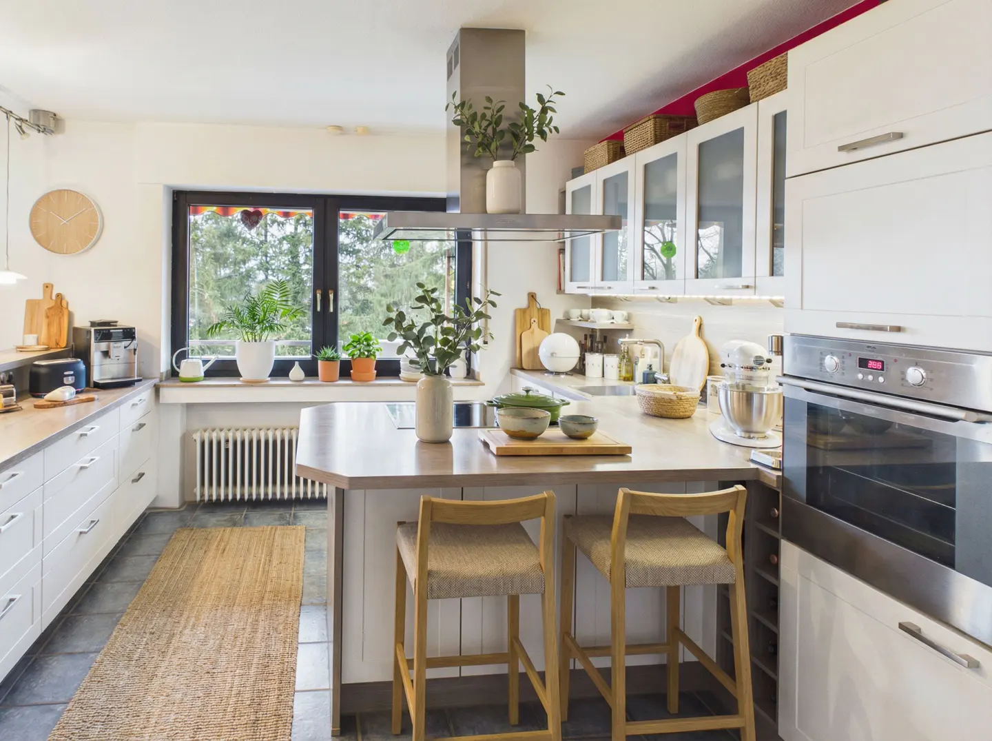 Bright kitchen with white cabinets, stainless steel appliances, and a center island with two wooden stools. A large window overlooks a green outdoor space.