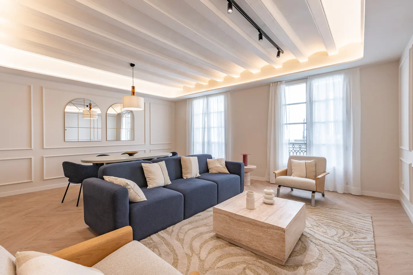 Bright living room with a blue sofa, beige rug, and light wood floors. Two arched mirrors hang above a dining table with chairs. Windows with white curtains let in natural light.