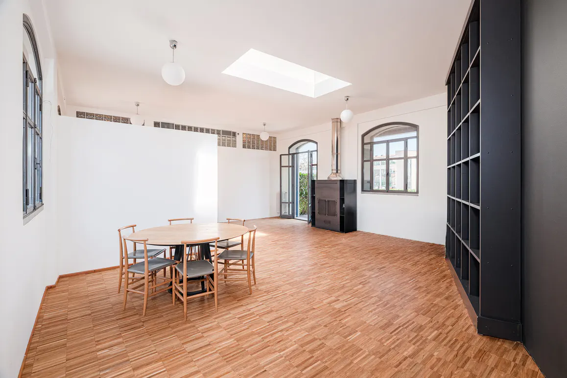 Bright, airy room with wood floors, white walls, and arched windows. A round table with chairs sits near a white partition. A black bookshelf and stove add contrast.