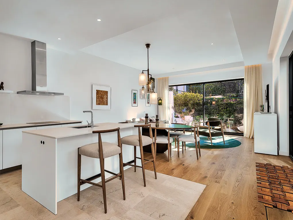 Bright, modern kitchen with white cabinets, island with stools, and stainless steel range hood. Dining area with table and chairs, view to garden.