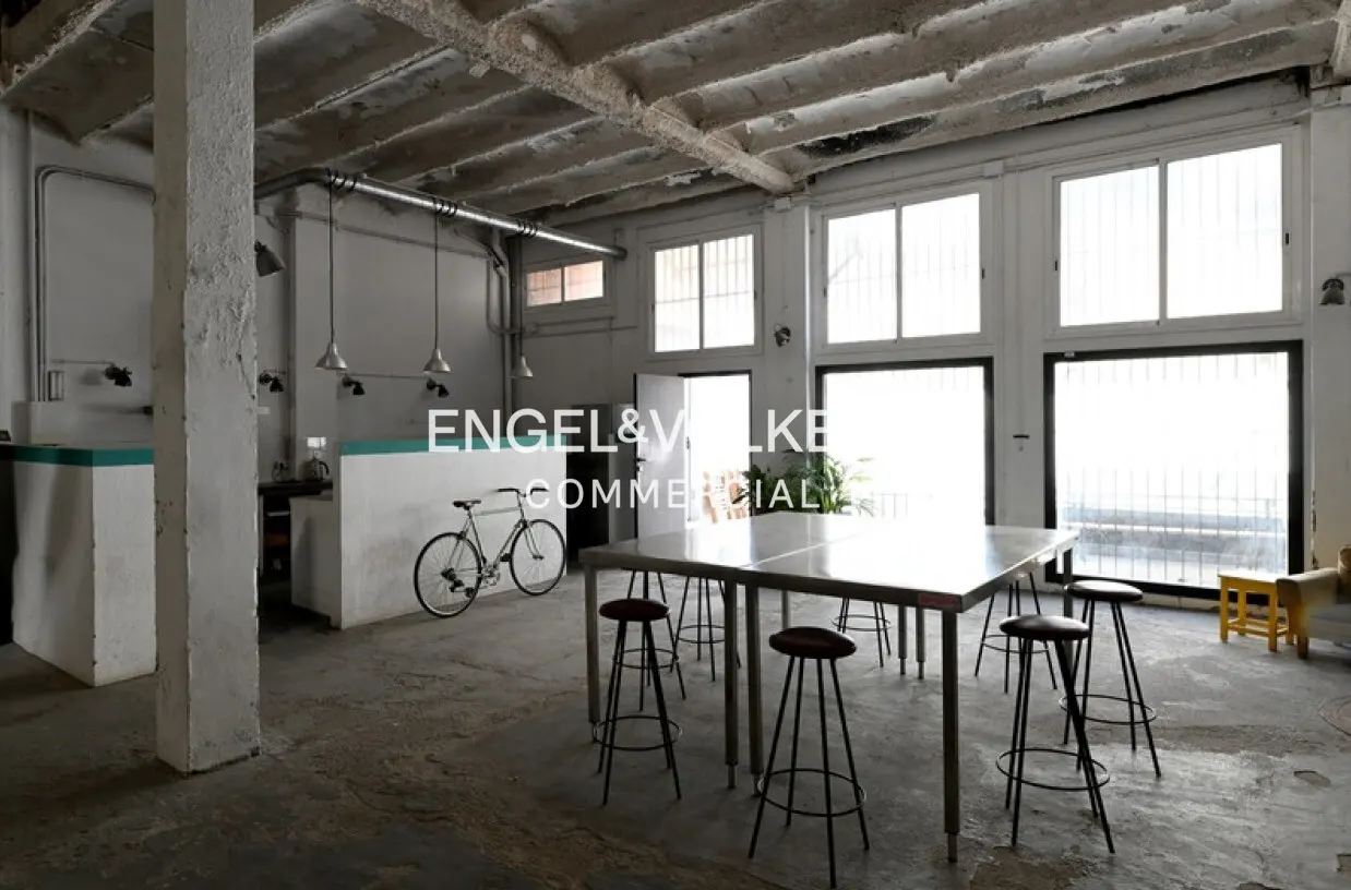 Industrial-style open space with concrete floors, high ceilings, a large metal table with stools, and a bicycle.