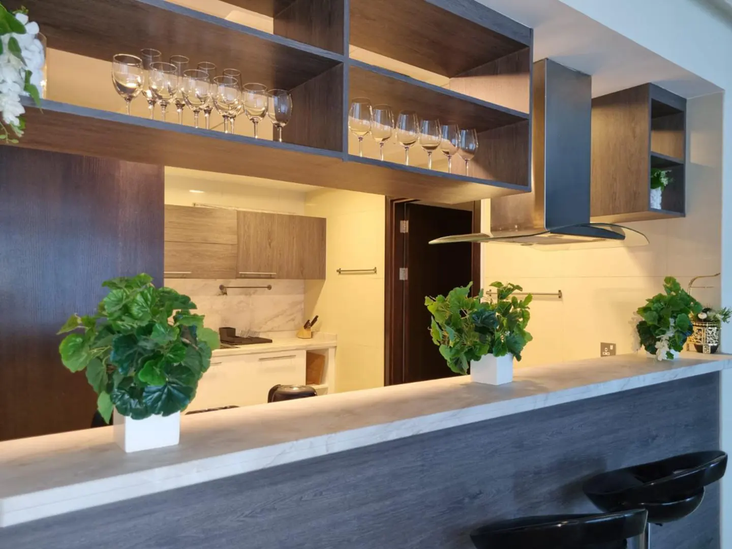 A modern kitchen with gray cabinets, open shelving displaying wine glasses, and a breakfast bar with black stools. Green plants add a touch of nature.