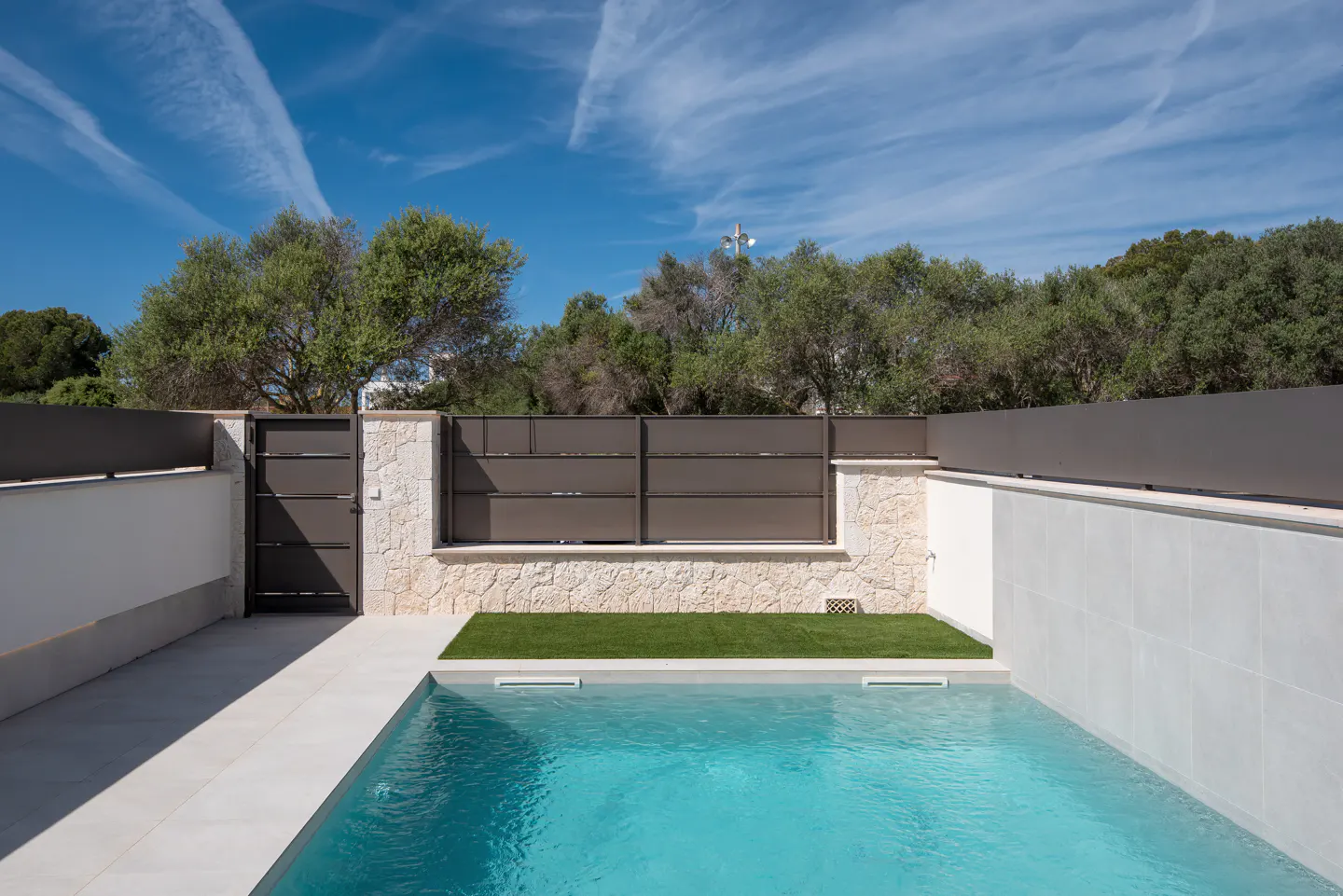 A backyard pool with turquoise water is surrounded by white tile and a stone and metal fence. Green trees and a blue sky are in the background.