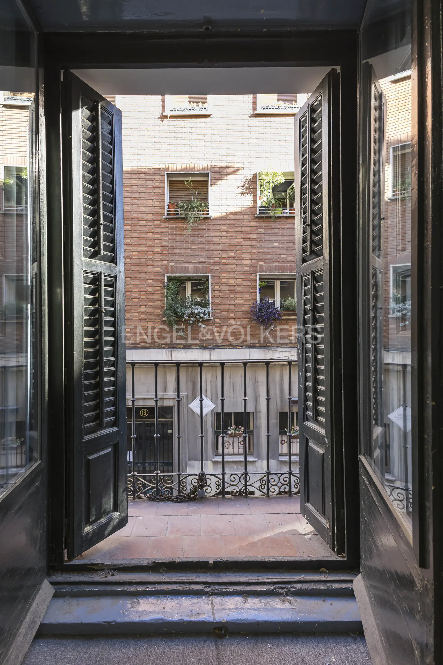 View from open window with black shutters, overlooking a brick building with flower boxes and an Engel & Völkers sign.