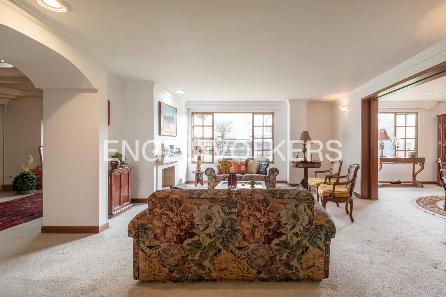 Spacious living room with floral sofa, chairs, and large windows. Neutral carpet and white walls create a bright, airy feel.