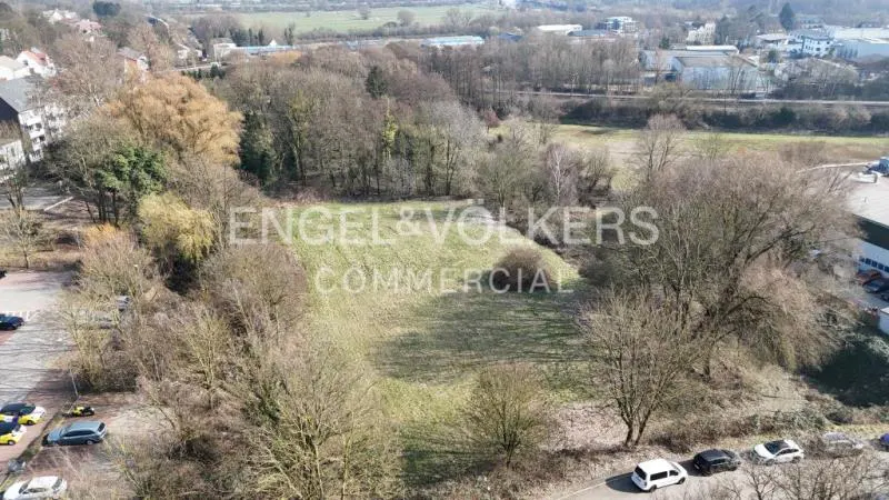 Aerial view of a grassy commercial lot surrounded by trees, with the Engel & Völkers logo superimposed. Cars are parked nearby.