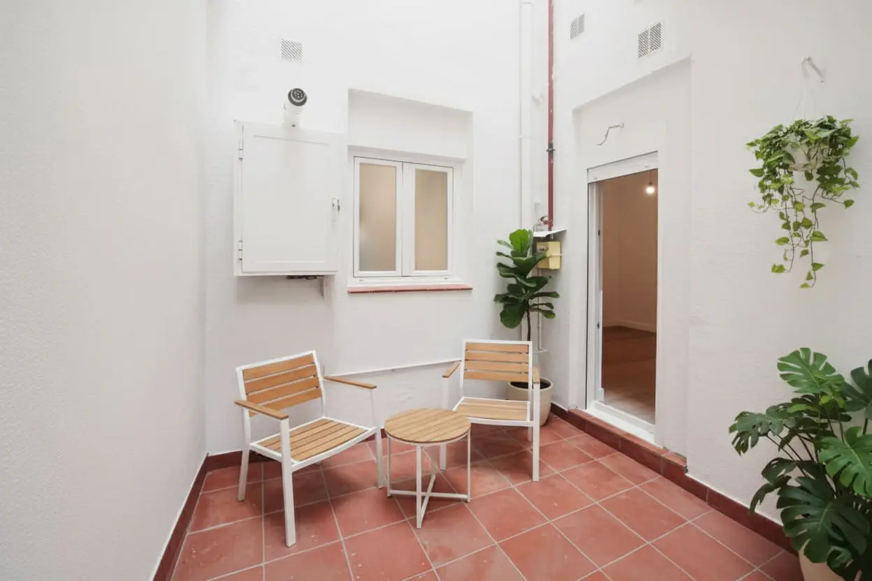 Bright patio with two wooden chairs, a small table, and plants on red tile floor. White walls and a window add to the airy feel.